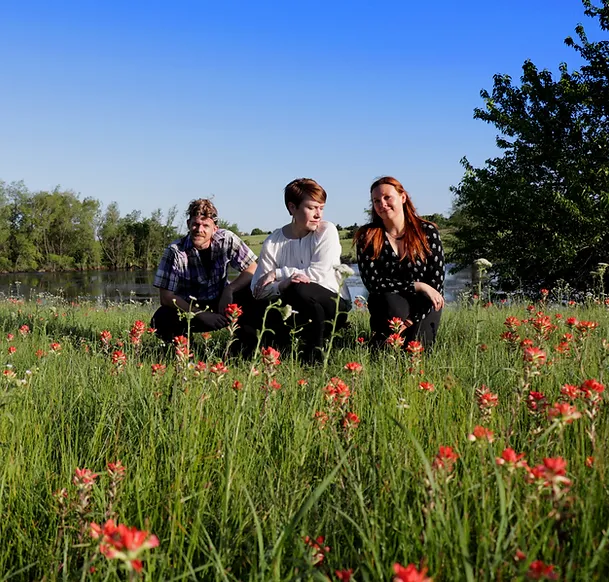 Three people squatting in a field by a pond.